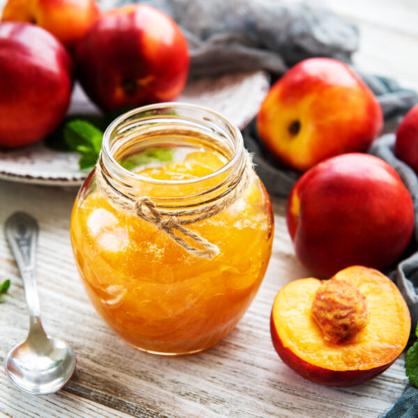 Löffel neben Glas mit Nektarinen-Marmelade in geöffnetem Gläschen steht neben frischen Nektarinen und Minze auf Tischdecke