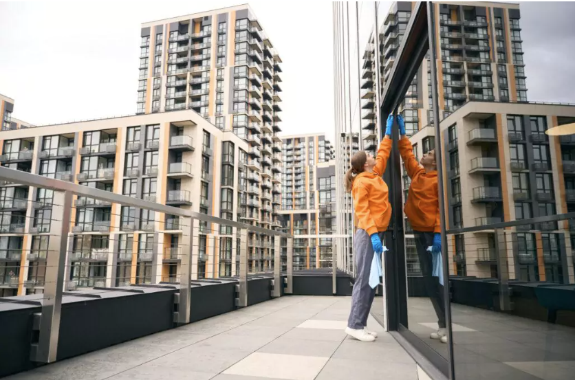 Frau mit oranger Jacke, grauer Hose und blauen Hygienehandschuhen steht vor einer Glasfront und putzt die Scheiben. Dahinter viele Hochhäuser zu sehen