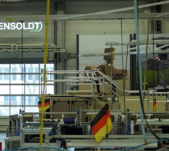 Einblick in eine Produktionshalle von HENSOLDT mit dem Firmenlogo im Hintergrund und der deutschen Fahne im Vordergrund