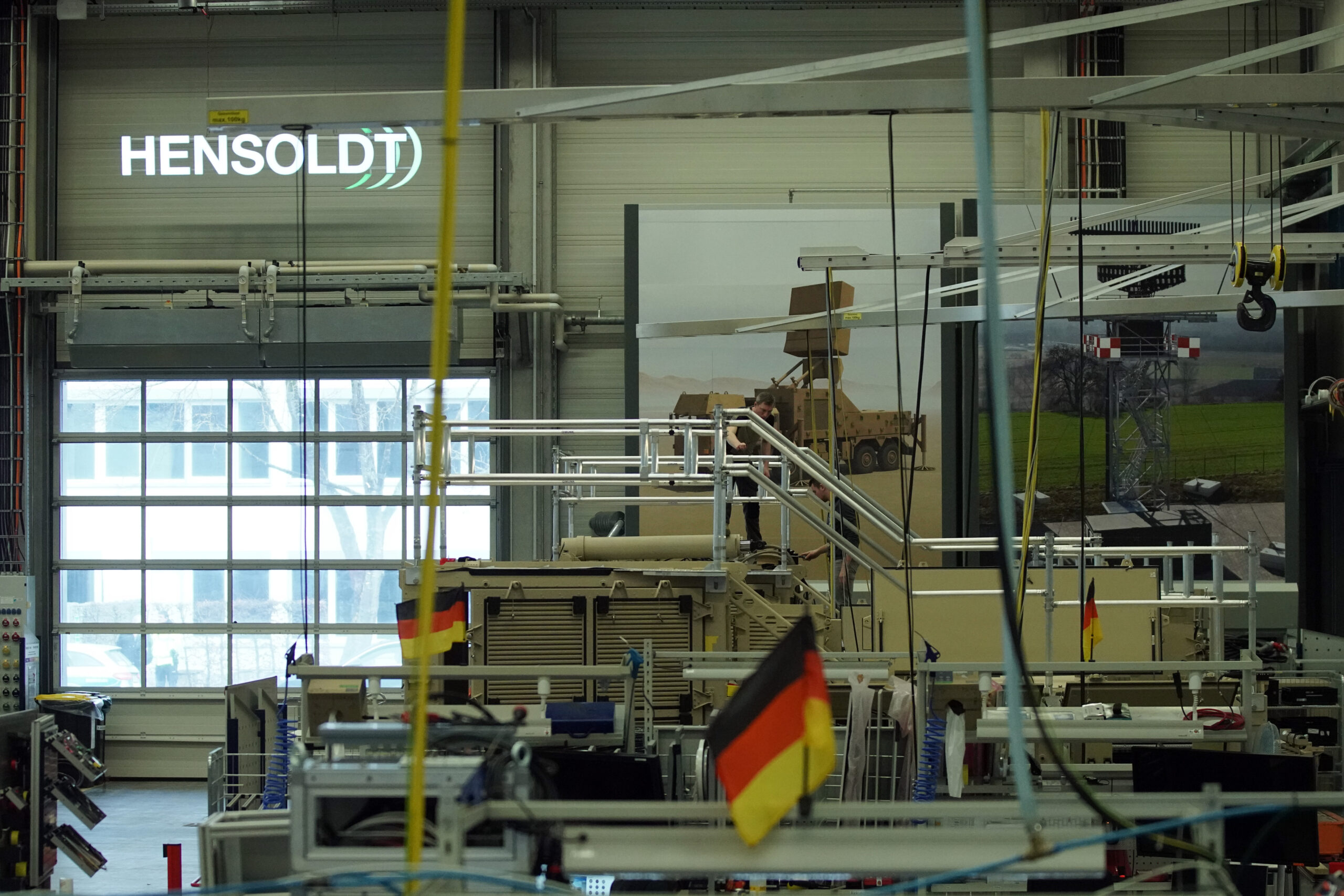 Einblick in eine Produktionshalle von HENSOLDT mit dem Firmenlogo im Hintergrund und der deutschen Fahne im Vordergrund